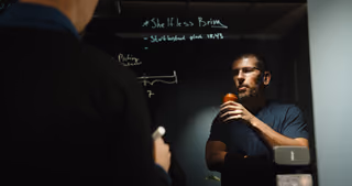 Colleagues discussing a problem on opposite sides of a glass whiteboard.